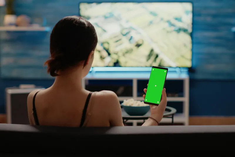 Caucasian woman holding mobile phone with green screen vertically looking at mockup template for chroma key. Young person using modern smartphone with copy space of isolated background